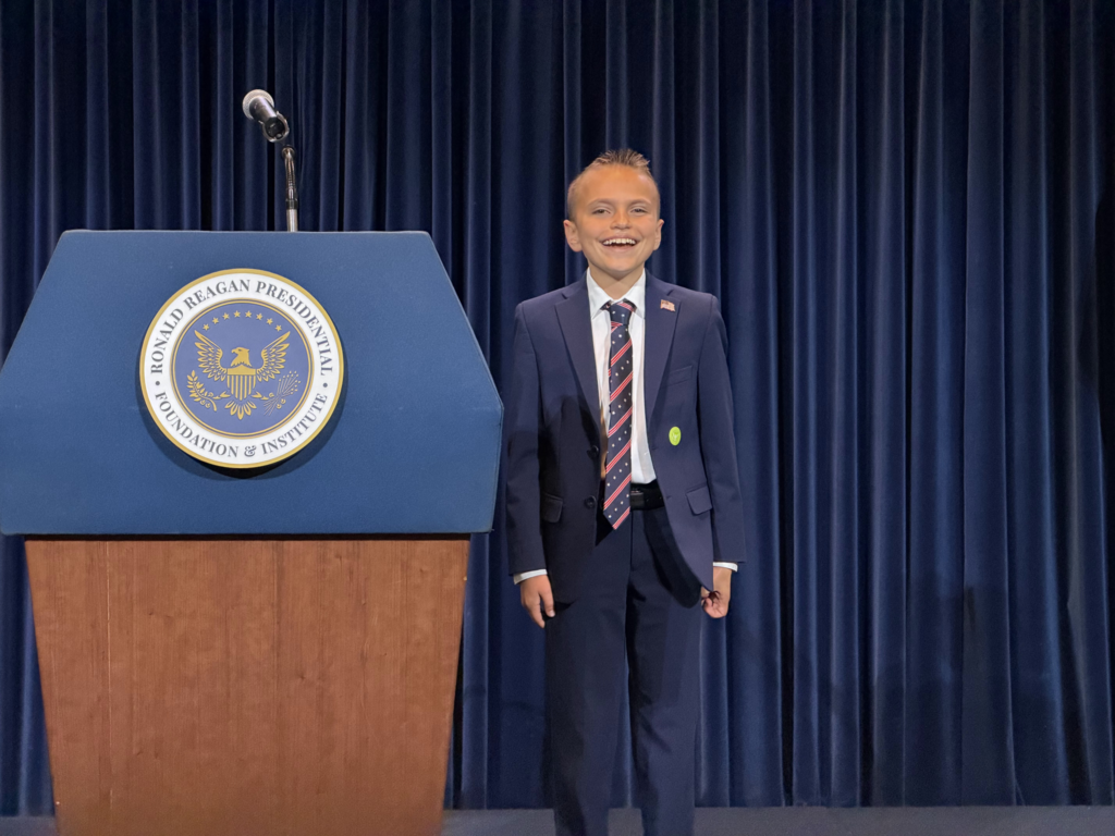 Logan dressed in a business suit stands next to a podium with a presidential seal
