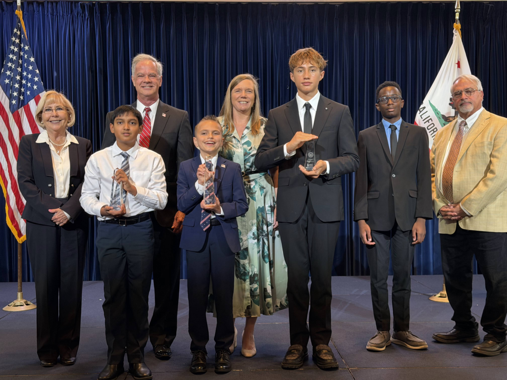 a group of adults stand on stage with student winners. The American flag on the left of the group