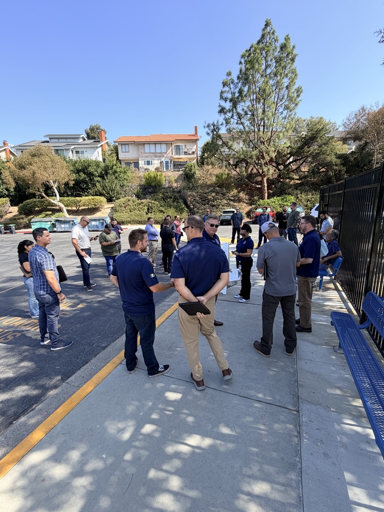 several people stand on a sidewalk near a black fence. Hills and homes in the background