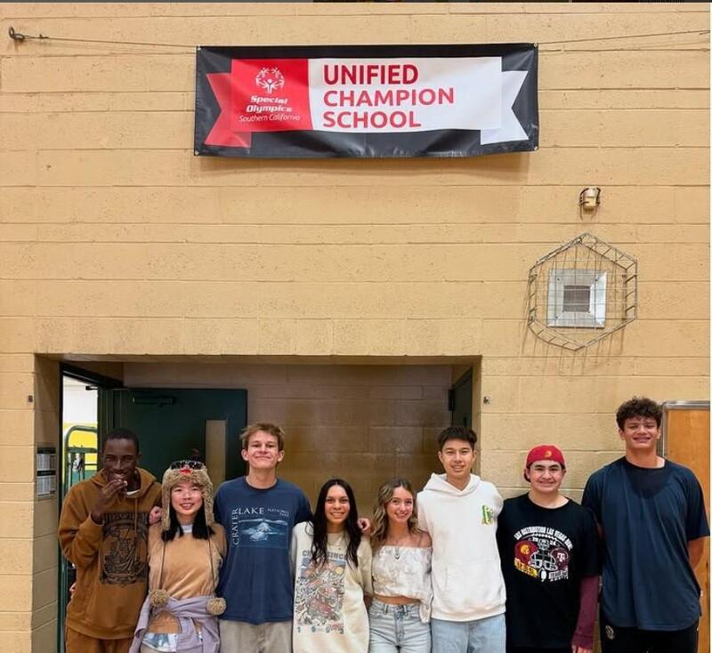 a group of students with a banner hung above on a biege wall. The banner is black, white and red with text "Unified Champion School"