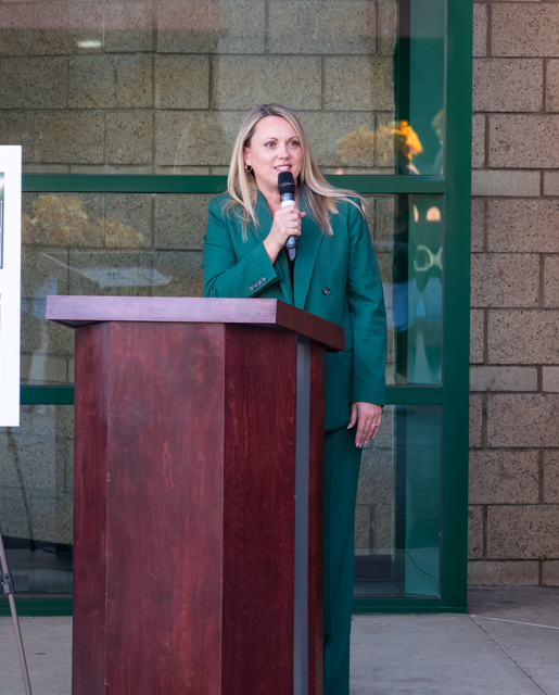 brinda leon holds a microphone and stands to right of a dark wood podium. Glass windows behind her with green painted edging