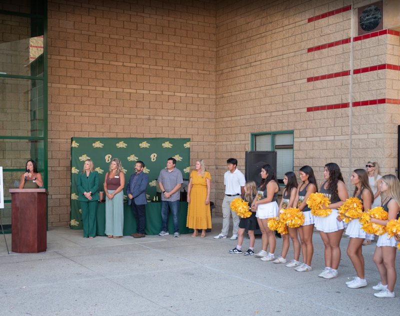 board president carrie flanders stands at podium with other members of board to the right and in front of a BOHS green and gold step and repeat backdrop. Other cheerleaders stand to the right of the board.