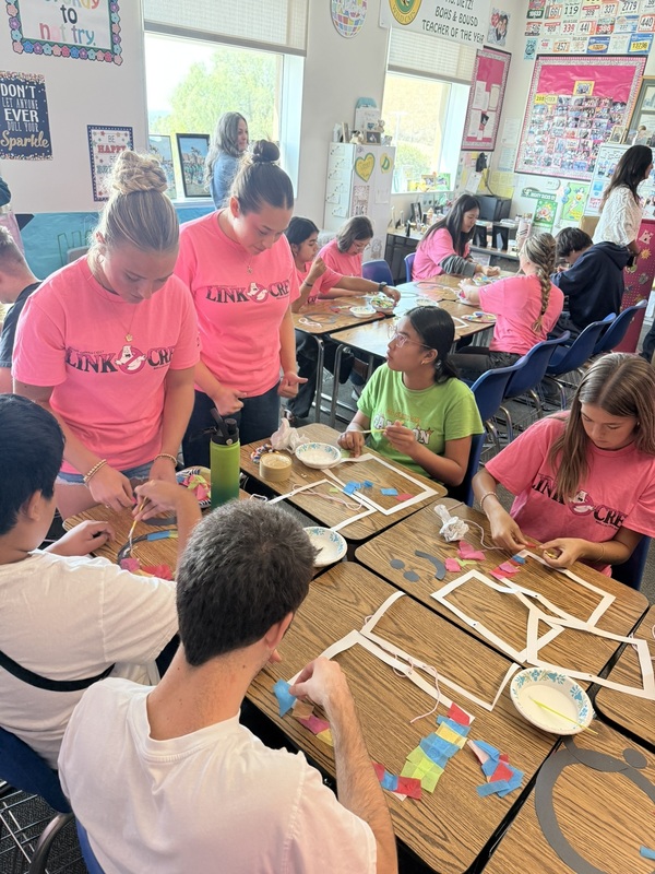 students with pink shirts stand over a table with craft and art supplies to help other students with a project