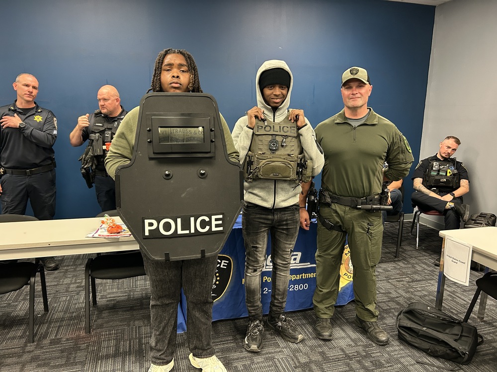 Two BHSD 228 students pose with a police officer at the Delta Academy Career Fair at South Suburban College. One holds a police shield, while the other wears a police tactical vest. Other officers and informational tables are visible behind them.