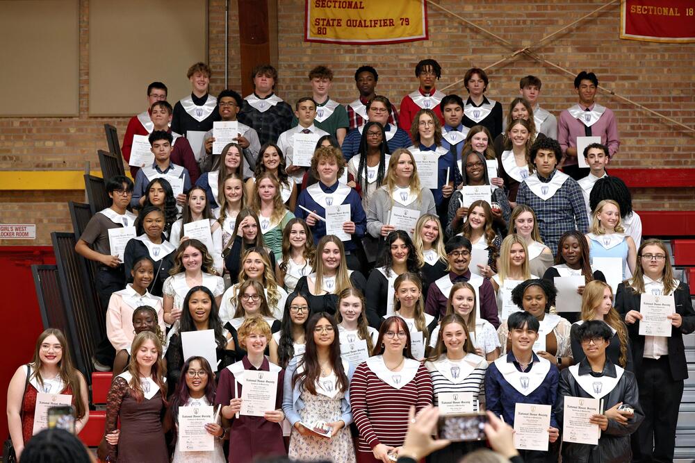 Tinley Park High School National Honor Society members pose in rows on gym bleachers, wearing white NHS stoles and holding membership certificates.