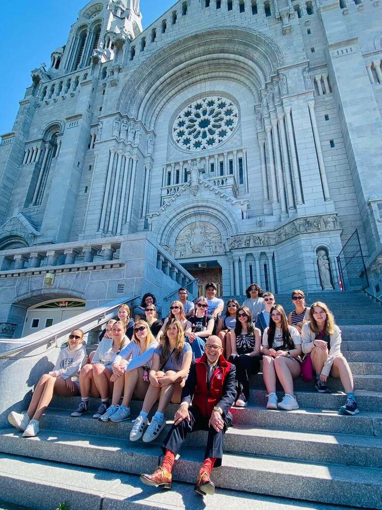 A group of Tinley Park High School students and chaperones sit on the steps of the Basilica of Sainte-Anne-de-Beaupré in Quebec, Canada, during a French language and cultural immersion trip. The students pose in front of the basilica’s grand stone façade, featuring ornate arches, columns, and a large rose window under a bright blue sky. The visit was one of many educational experiences that allowed students to explore the history, culture, and architecture of French-speaking Canada.