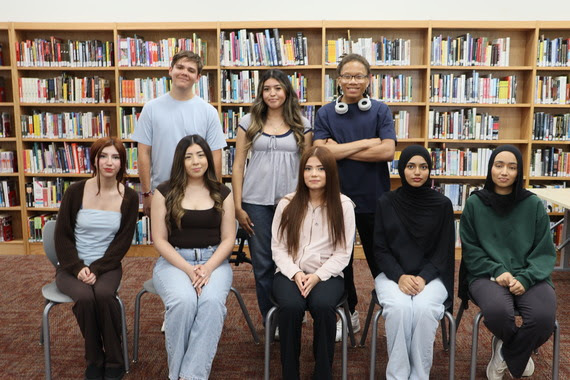 Eight students pose in two rows inside a school library, with bookshelves in the background.