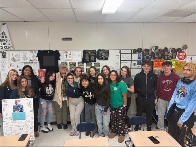 A group of high school students and an adult stand together in a classroom, smiling and posing for a photo. Posters, student artwork, and signs fill the walls behind them, and desks are visible in the foreground. One student holds a poster covered in handwritten messages, suggesting a supportive or celebratory gathering.