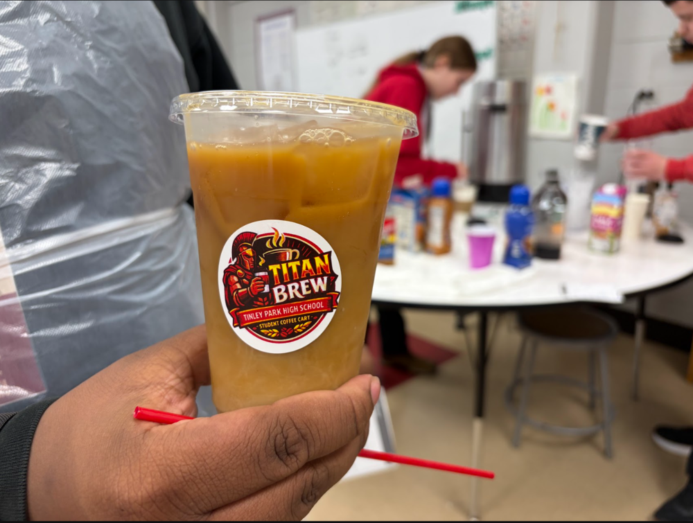 A person holds an iced coffee with a “Titan Brew—Tinley Park High School Student Coffee Cart” sticker while students prepare drinks at a table in the background.