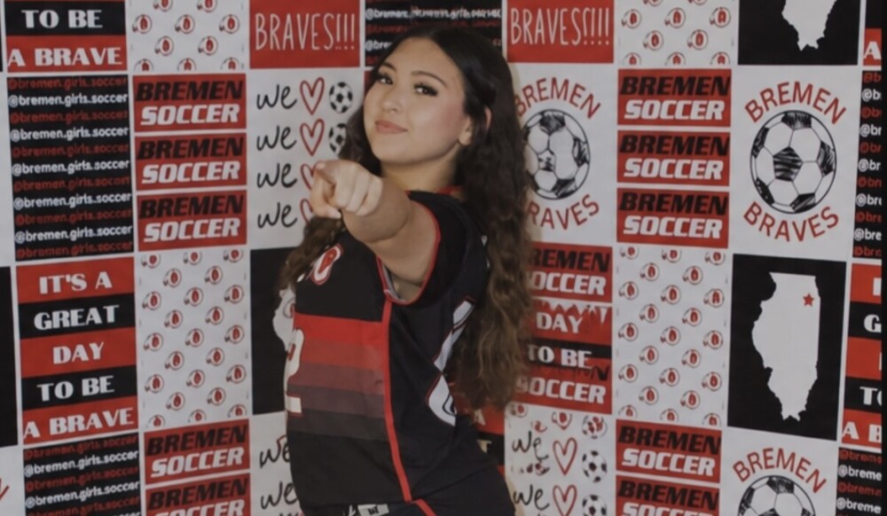 Sophia Lara poses in a Bremen Braves soccer jersey in front of a red, black, and white Bremen Soccer backdrop featuring soccer graphics and school spirit slogans. She points confidently toward the camera while smiling.