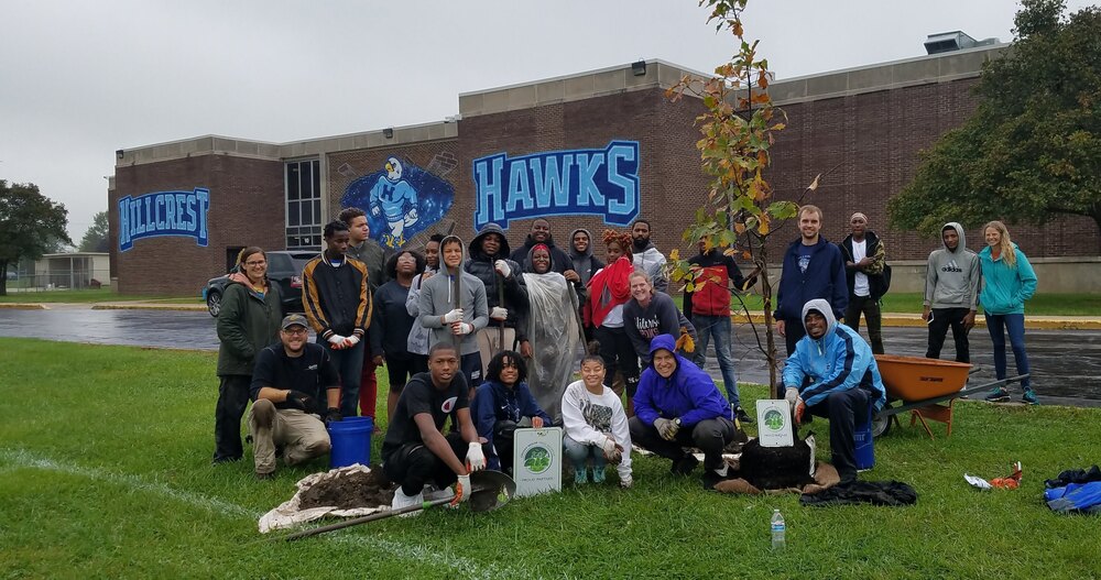 A group of Hillcrest High School students and staff pose in rain gear beside a newly planted tree outside the school. Shovels, buckets and a wheelbarrow surround the group, with a large Hillcrest Hawks mural visible on the building behind them.