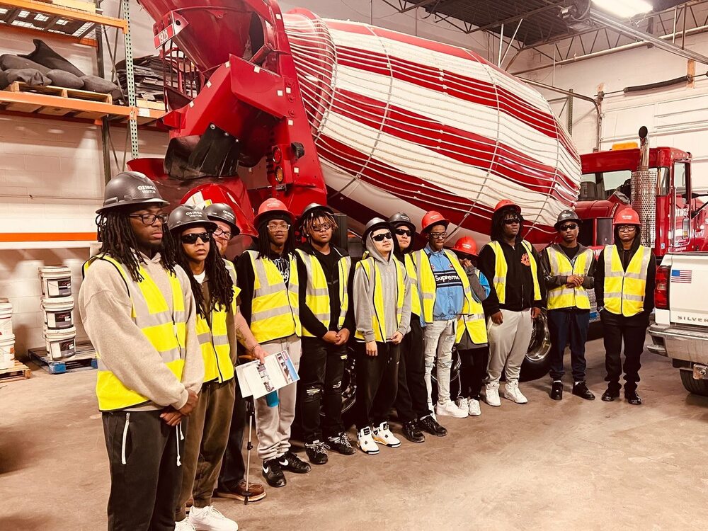 A group of students wearing hard hats, safety glasses and reflective vests stands in front of a red-and-white concrete mixer truck during a facility tour.
