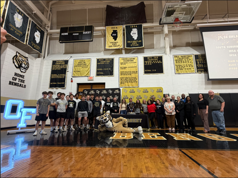 Group photo inside the Oak Forest High School gymnasium during the athletic banner unveiling ceremony. Football players, girls wrestling athletes, coaches, administrators, board members, and family members stand together on the basketball court beneath newly unveiled IHSA banners honoring the football team’s 2025 State Semifinal appearance and the girls wrestling team’s 2026 third-place state finish. Championship banners hang throughout the gym, with the school mascot posed in front of the group.