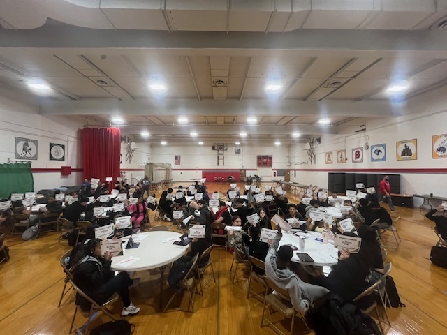 Students sit at round tables inside a high school gymnasium, holding up papers during a Junior Achievement JA Town financial literacy simulation. The gym features a wooden floor, high ceilings with bright lights, school banners on the walls, and a stage with red curtains in the background.