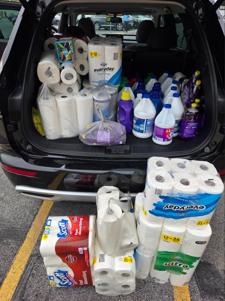 An open SUV trunk filled with paper towels, laundry detergent, bleach and cleaning supplies collected by Hillcrest High School’s Key Club for ANEW Domestic Violence Shelter, with additional packages of paper towels stacked behind the vehicle.