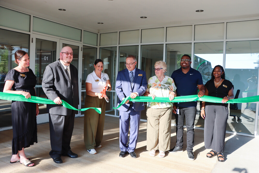 **Alt text:** Seven individuals stand outside the Bremen High School District 228 Administration Center holding a green ribbon during the official ribbon-cutting ceremony. The group poses in front of the building's glass entrance, with one person holding ceremonial scissors as they celebrate the opening of the District's new administrative offices and first-ever board room.