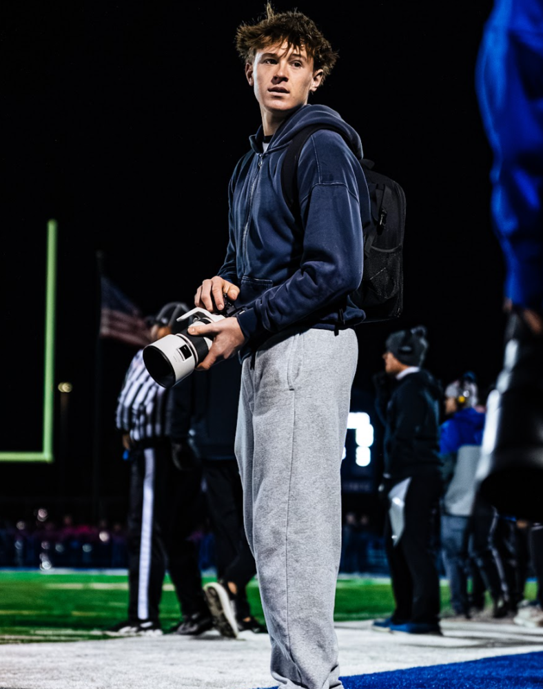 Nathan Dunaj of Oak Forest High School stands on the sideline of a football field at night, holding a camera with a long lens. He wears a navy hoodie, gray sweatpants, and a backpack, looking off to the side as players and coaches gather in the background under stadium lights.