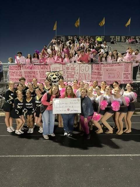 Oak Forest High School students, cheerleaders, staff and community members, many dressed in pink, gather on the football field with the Bengal mascot. People at the front hold a $2,132 check for the American Cancer Society, while pink breast cancer awareness banners hang behind the group.