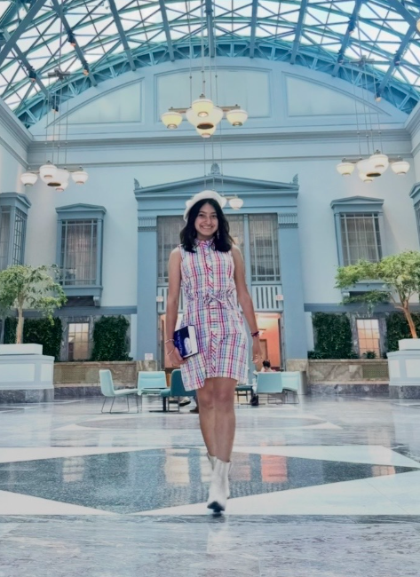 A young woman wearing a sleeveless plaid dress, white boots, and a white hat walks confidently through a bright indoor atrium with high glass ceilings, hanging lights, and elegant blue-and-white architecture. She is smiling and holding a small book or notebook at her side.