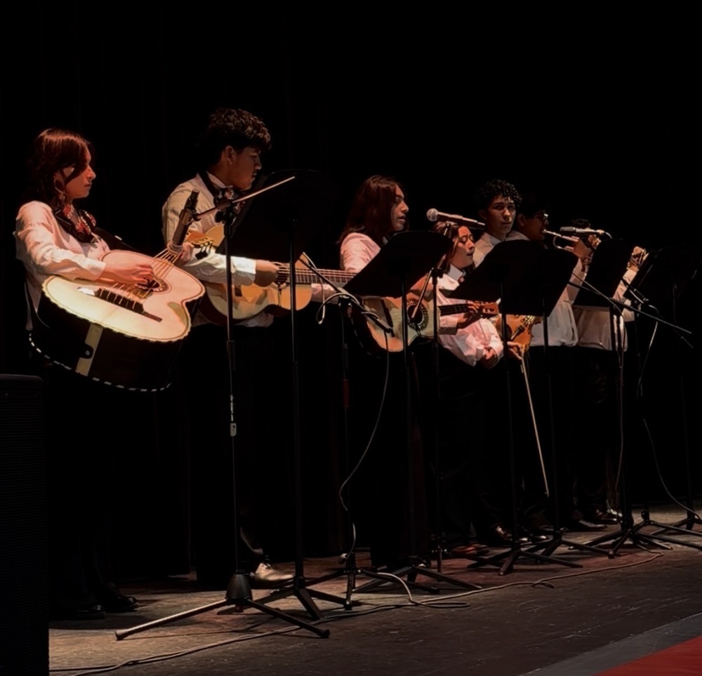 Bremen High School’s student mariachi ensemble performs onstage with string instruments and trumpets during the Hispanic Heritage Homeroom Takeover. Students wear white shirts and stand behind microphones and music stands.