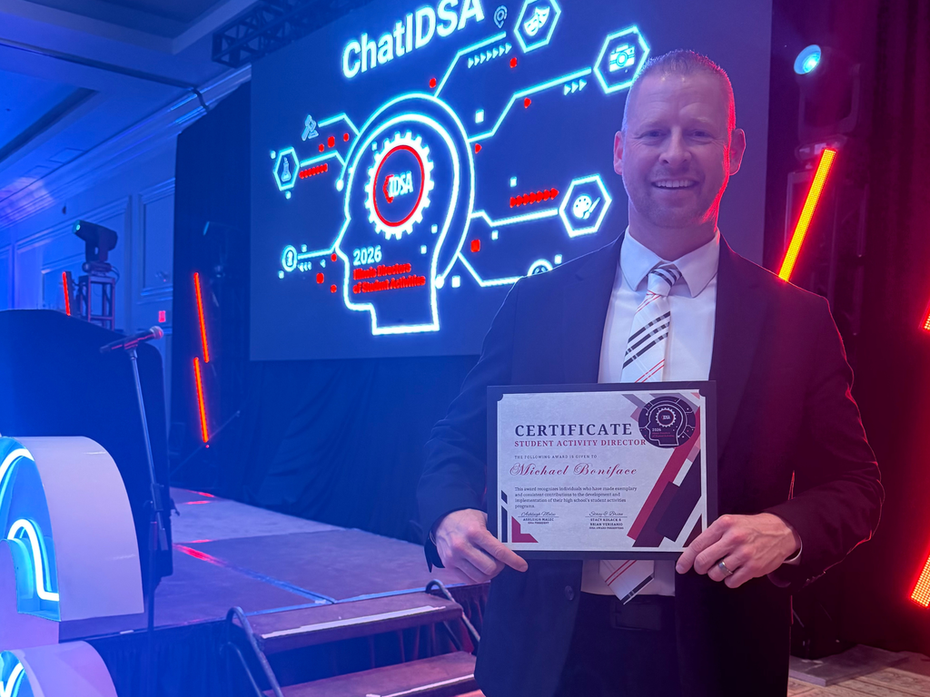 Dr. Michael Boniface stands on stage at the 2026 Illinois Directors of Student Activities (IDSA) Awards Banquet, smiling and holding his Student Activity Director Recognition Award certificate. Behind him is a large illuminated screen displaying “ChatIDSA” and a graphic design with the IDSA logo.