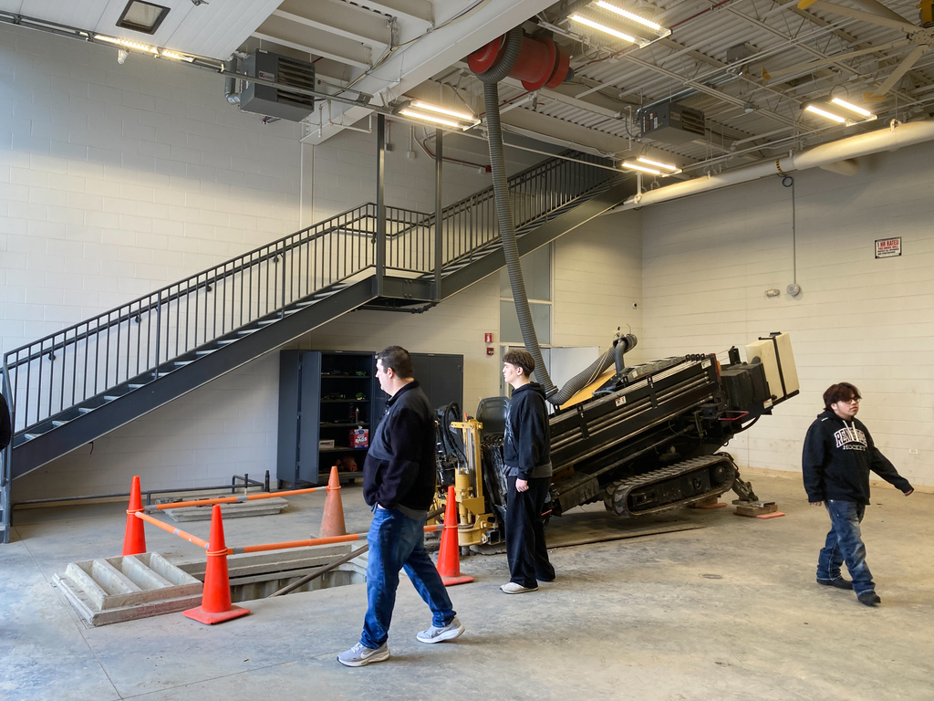 Students and an instructor walk through an indoor training area featuring drilling equipment, safety cones, and a staircase leading to an upper level.
