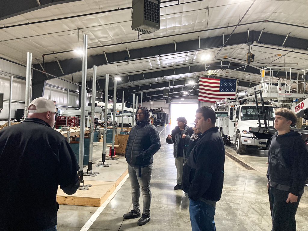 A group of students listens to an instructor inside a large equipment garage with utility trucks and an American flag hanging in the background.