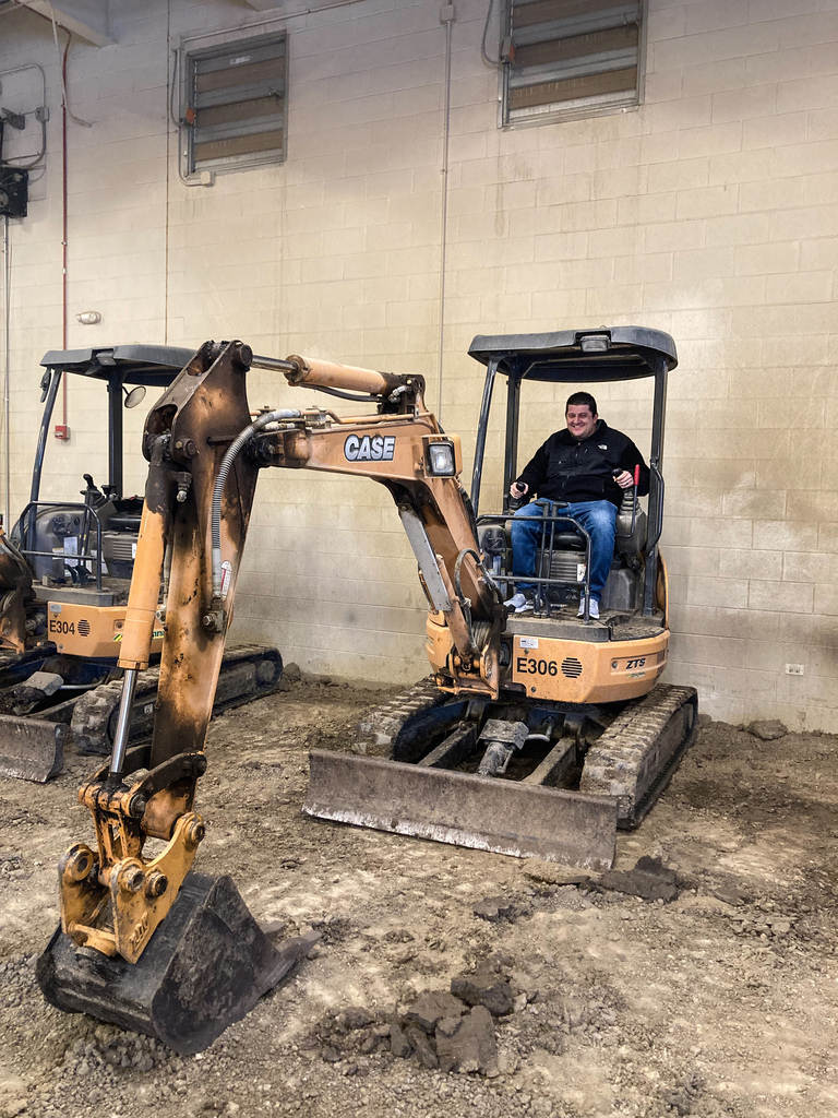 A student smiles while operating a small CASE mini excavator inside a training garage filled with dirt and heavy equipment.