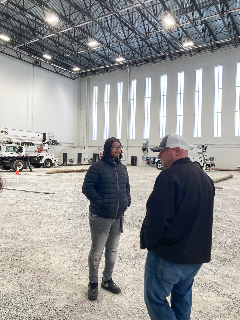 A student speaks with an instructor inside a large indoor training facility with high ceilings, utility trucks, and wooden utility poles set up for practice.
