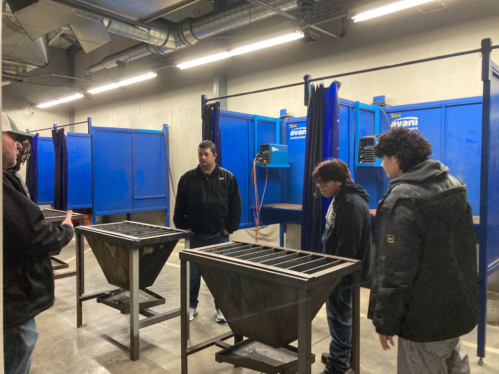 Students observe metal fabrication projects inside a welding lab with blue welding booths and training stations lining the walls.