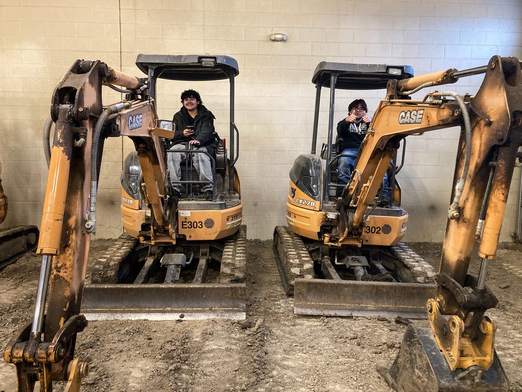 Two students sit in separate CASE mini excavators parked side by side inside a training space, smiling and posing while seated at the controls