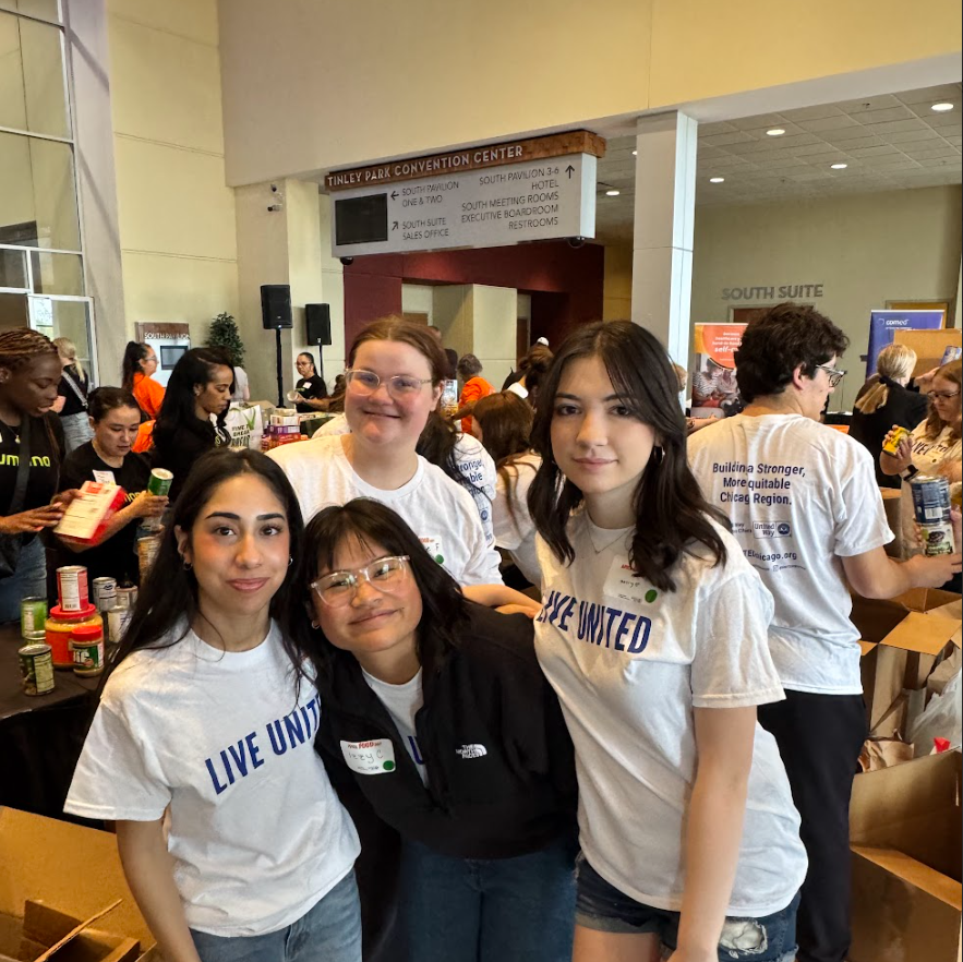 Four high school students wearing white “Live United” T-shirts pose together inside the Tinley Park Convention Center, smiling at the camera. Behind them, other volunteers sort and pack food items at tables filled with canned goods and boxes, with signage for the convention center visible overhead.