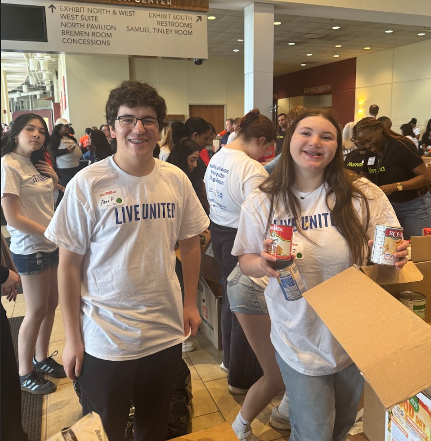 Two high school students wearing white “Live United” T-shirts smile at the camera while volunteering inside the Tinley Park Convention Center. One student holds canned food items while standing next to an open box, as other volunteers in the background sort and pack donations at tables.
