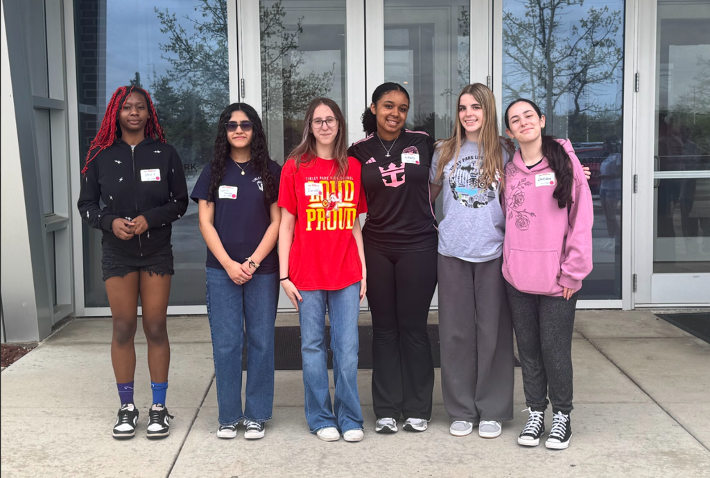 Six high school students stand side by side outside a building entrance with glass doors, posing for a photo. They wear casual clothing, including T-shirts, hoodies, and sneakers, and have name tags on their shirts. The group smiles at the camera, standing close together on the sidewalk.
