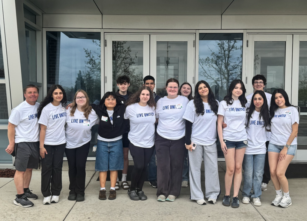 A group of high school students and one adult stand together outside a building entrance with glass doors, posing for a photo. Many are wearing white “Live United” T-shirts, standing shoulder to shoulder and smiling at the camera.