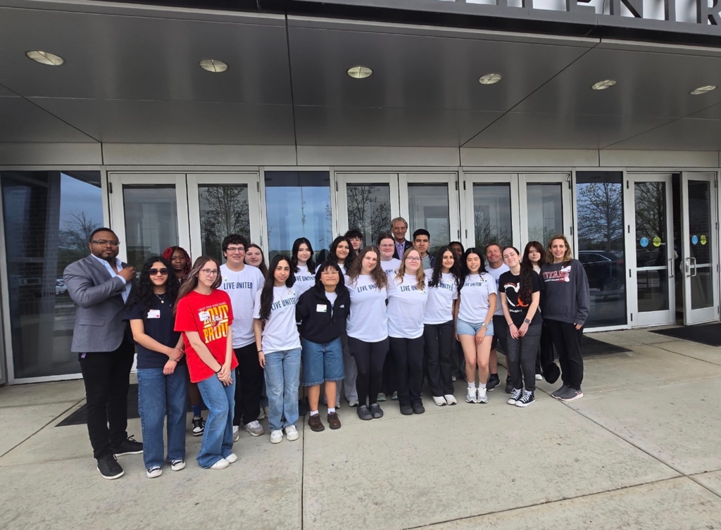 A larger group of high school students from Oak Forest and Tinley Park stand together outside a building entrance with glass doors, joined by a few adults. Many students are wearing white “Live United” T-shirts, while others wear casual clothing. The group faces the camera, smiling, with the building’s overhang and entryway visible above them.