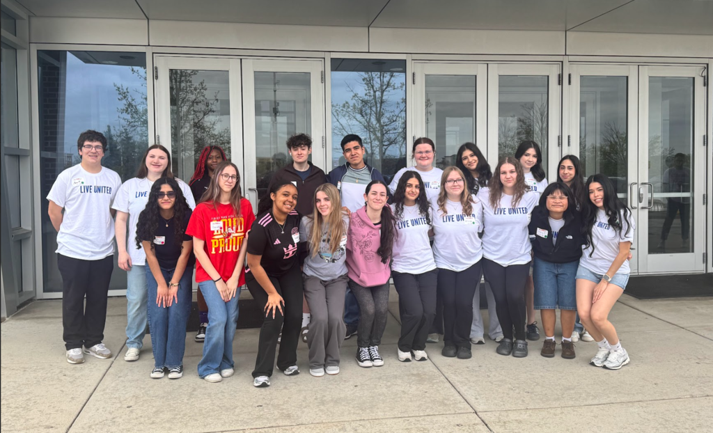 A group of high school students from Oak Forest and Tinley Park pose together outside a building entrance with glass doors. Many are wearing white “Live United” T-shirts, while others wear casual clothing. The students stand closely together, smiling at the camera, with some crouching slightly in the front row.