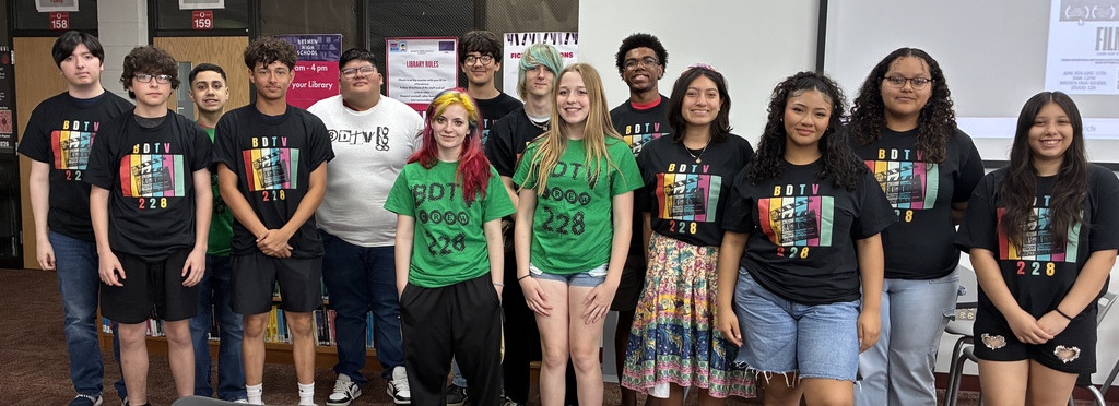 **Alt text:** A group of 15 Bremen High School District 228 students who participated in the BDTV Summer Camp pose together in a school library. The students wear black or green BDTV 228 and BDTV Crew T-shirts, standing in two rows and smiling at the camera. Bookshelves, library signage, and a presentation screen are visible in the background, highlighting the media-focused summer learning environment.