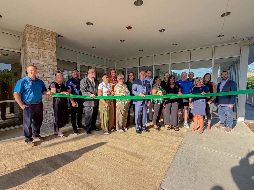 **Alt text:** A group of District 228 Board of Education members, administrators, staff, community leaders, and guests stand outside the Bremen High School District 228 Administration Center holding a green ribbon during the official ribbon-cutting ceremony. The group poses in front of the building's entrance in celebration of the opening of the District's new administrative offices and first-ever board room.