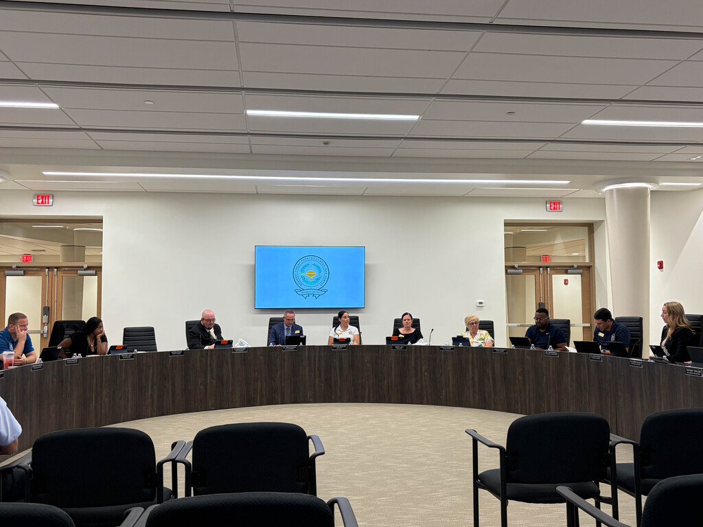 **Alt text:** Members of the Bremen High School District 228 Board of Education and district leadership are seated around the new board room dais during a board meeting. The modern meeting space features a large digital display with the District 228 logo and audience seating in the foreground, highlighting the District's first-ever dedicated board room.