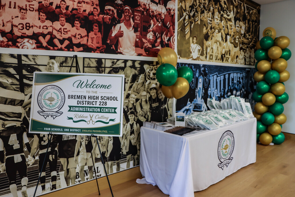 **Alt text:** A welcome display for the Bremen High School District 228 Administration Center ribbon-cutting ceremony features a welcome sign on an easel, a registration table with District-branded materials, and green and gold balloon columns. The display is set against a large wall mural showcasing historic photos of District 228 students, athletes, and school pride.