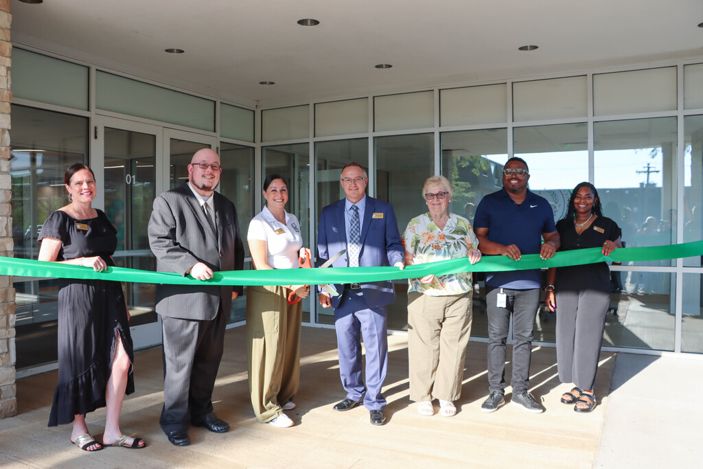**Alt text:** Seven individuals stand outside the Bremen High School District 228 Administration Center holding a green ribbon during the official ribbon-cutting ceremony. The group poses in front of the building's glass entrance, with one person holding ceremonial scissors as they celebrate the opening of the District's new administrative offices and first-ever board room.