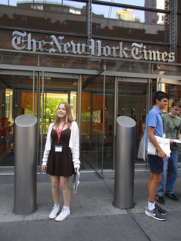 **Alt text:** Margaux, a student from Tinley Park High School, smiles while standing outside The New York Times headquarters during a journalism internship visit. Wearing a black dress, white cardigan, and a red internship lanyard, she holds newspapers in front of the building's glass entrance as other student interns stand nearby.