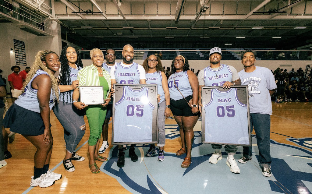 **Alt text:** A group of nine people stands together at center court in a high school gymnasium during a special recognition ceremony at Hillcrest High School. Two framed basketball jerseys reading "MAMA BA 05" and "MAC 05" are displayed in front of the group, while another person holds a commemorative plaque. Several members wear gray Hillcrest basketball jerseys, and the gym is filled with spectators in the background as the honorees pose for a celebratory photo.