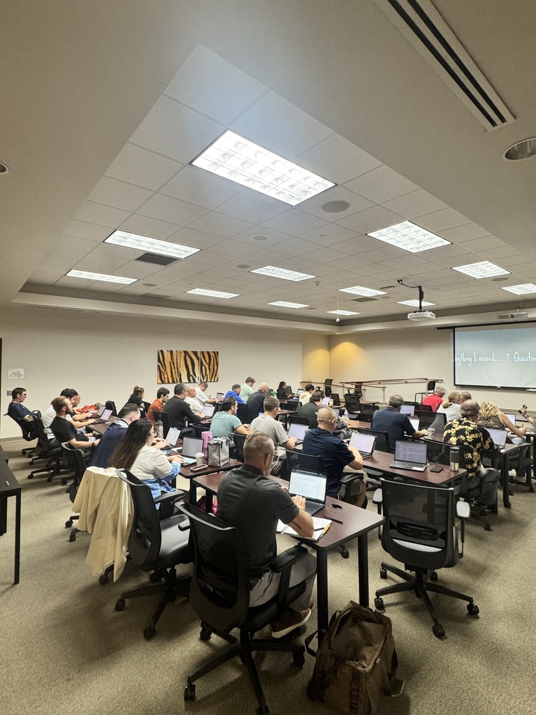 Staff members work on laptops during a professional development session in a conference room while a presentation is displayed at the front.