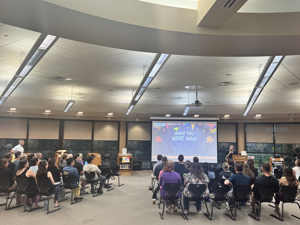 Staff members attend a presentation in a school library or media center, facing a screen displaying updates and reminders for the new school year.