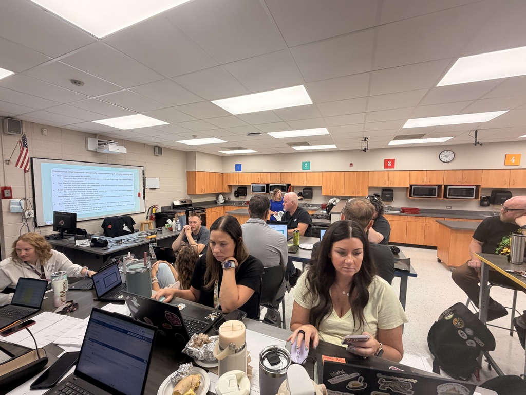 Staff members collaborate in small groups with laptops and notes during a professional development session in a classroom.