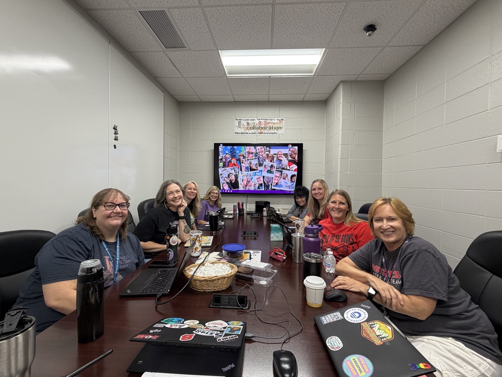 Eight staff members smile around a conference table during a collaborative team meeting, with laptops, drinks and work materials spread across the table.