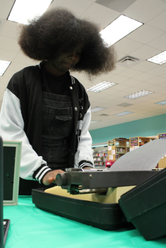 Caption: Writing Like It’s 1984: Senior Arielle Wright tries writing on a 1984 Olympia International typewriter. “ I know essentially how they work. It’s stiff and you had to press really hard for it to work. I prefer keyboards, they’re faster to type with. I guess that’s why people typed so slowly back then!” said Wright.  Part of the Media Center Dedication Anniversary celebration featured a Then and Now table, where vintage items like a 1962 Kodak film projector and the typewriter were displayed next to their modern replacements. Students had the opportunity to touch and interact with the items and learned how far our technology has come. “Students back then would probably think today’s high-tech media center would be odd,” Wright said.  Photo by Kyle McLaren