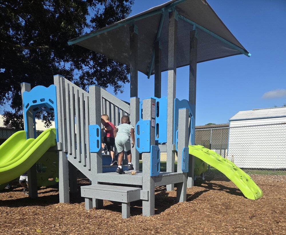 Students playing on a new grey, blue, and lime green playground structure set over fresh woodchips on a sunny day at Columbia Elementary. The photo is taken from behind so no student faces are visible.